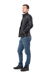 Wall Mural - Side view of young casual man with hands in leather jacket pockets looking up serious. Full body length portrait isolated over white background.