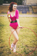 Beautiful brunette woman in pink retro body outfit with ponytail hairstyle, hula hoop and soda bottle in her hand. Outdoors