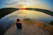 © raisondtre - Young woman sitting on the stone enjoying peaceful moment of sunset. In the reflection of the lake water sees clouds and sun.