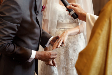 Priest holds microphone while groom puts wedding ring on brides