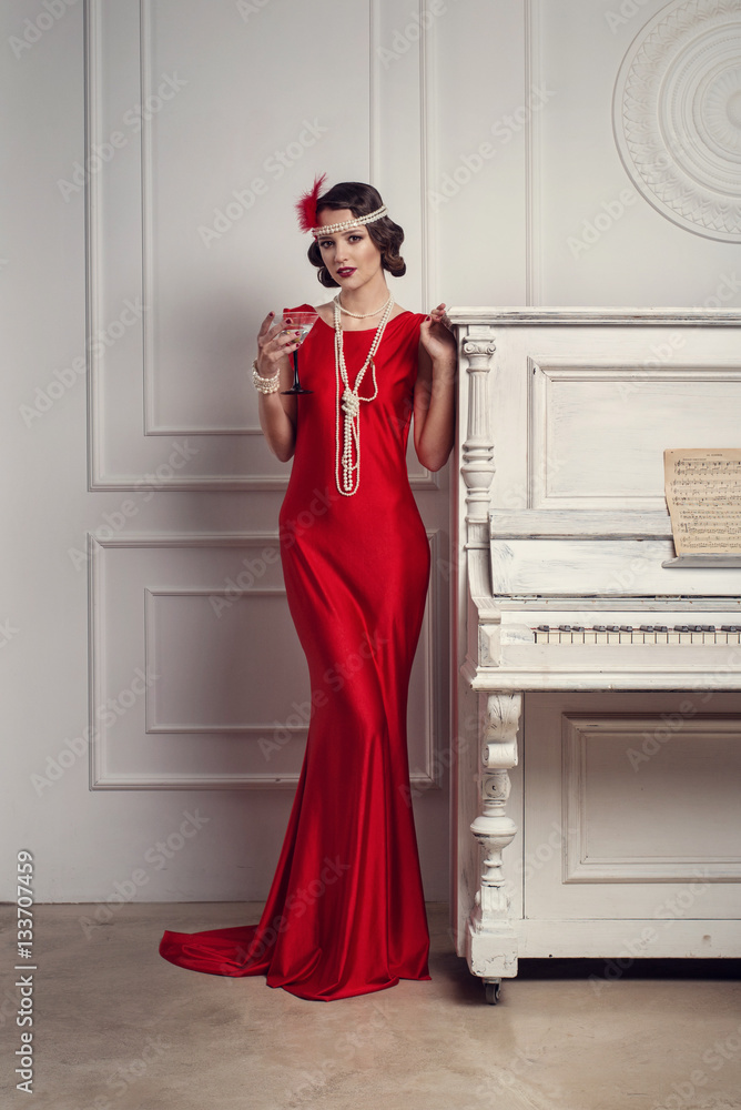 Young beautiful girl in red dress style of the 20's or 30's with glass of  martini near the piano. Vintage style beautiful woman. Old fashioned makeup  and retro finger wave hairstyle. Stock