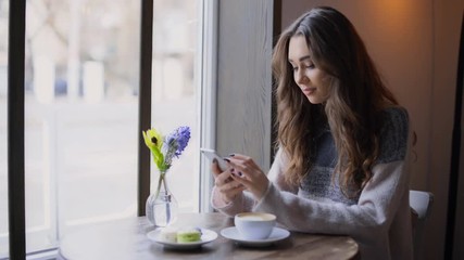 Wall Mural - Beautiful young woman sitting at cafe and waiting for her boyfriend while texting on mobile phone