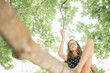 © Mint Images - A young girl in shorts sitting on a high tree branch under a canopy of green leaves.