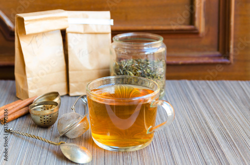 Fotografia, Obraz Glass cup of hot tea with spoon infuser and tea jar on grey straw background