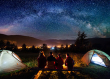 Silhouette Of Four People Sitting On A Bench Made Of Logs And Watching Fire Together Beside Camp And Tents In The Night. On The Background Starry Sky, Milky Way, Mountains And Luminous Town. Rear View