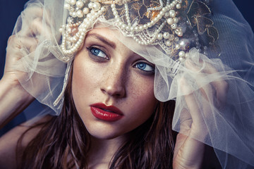 fashion beauty portrait of young beautiful young woman with makeup and freckles on her face and pearl headpiece on her head and white tulle in front of her face on dark blue background. 
