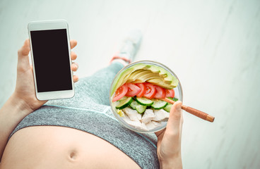 Wall Mural - Young woman is eating a salad  and using a fitness app on her smartphone after a workout.