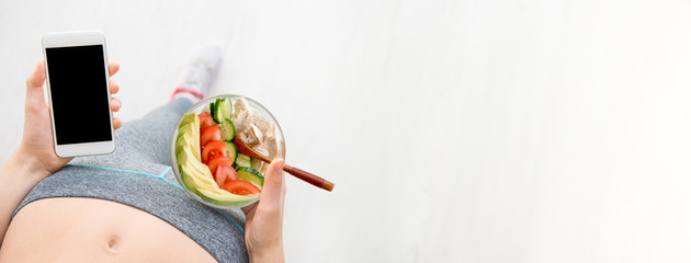 Wall Mural - Young woman is eating a salad  and using a fitness app on her smartphone after a workout.