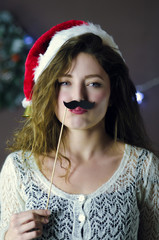 Blond curly beautiful smiling girl wearing Christmas hat holding fun paper mustache on Christmas decorations background