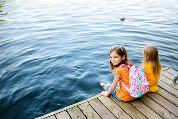 Wall Mural - Two cute little girls sitting on a wooden platform by the river or lake