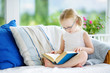 © MNStudio - Adorable little girl wearing eyeglasses reading a book in white living room