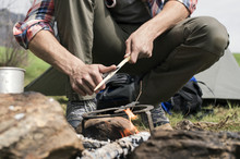 Low Section Of Man Cutting Stick Over Bonfire At Campsite