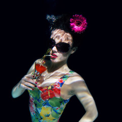 beautiful girl in a colorful stylish swimsuit and sunglasses and pink gerbera in the head with a cocktail  in her hand underwater in the swimming pool