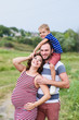 © o_lypa - Pregnant family of three in expectation of baby. Father mother and son playing together in day outside. Happy people hugging. Smiling laughing baby sitting on shoulders of daddy.