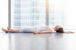 © fizkes - Young woman practicing yoga, lying in Savasana exercise, Dead Body, Corpse pose, working out, wearing sportswear, white t-shirt, pants, indoor full length, near floor window with city view, side view