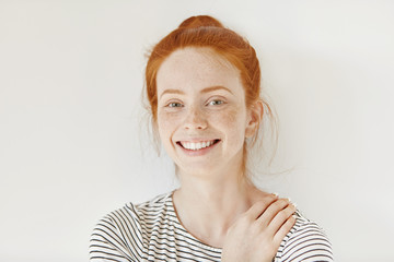 Stylish freckled teenage girl with hair knot having fun indoors and looking at camera with cheerful smile. Beautiful young redhead lady smiling happily keeping hand on her breast, isolated on white