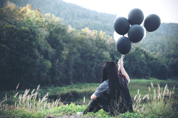 A turn back asian woman holding black and white balloons while sitting with green mountain and lake background