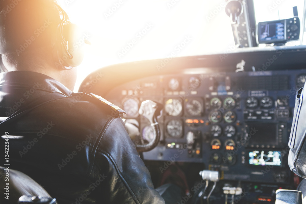 Adult airplane pilot wearing headset and outfit performing his job, sitting inside aircraft cockpit at steering control with modern dashboard. Bright sunshine penetrating into cabin through glass - obrazy, fototapety, plakaty 