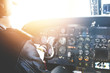 Caucasian aviator piloting airplane. Rear view of commercial pilot wearing headset and leather jacket sitting inside cockpit in front of dashboard with numerous buttons. Blurred shot, flare sun