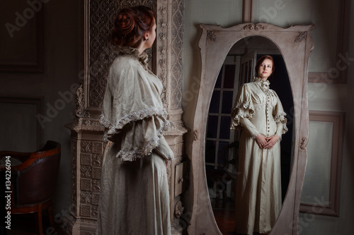 Plakat Kobieta w stylu retro szlachcianka stoi przed lustrem próbuje na sukienkę.