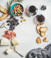 Wine and snack set. Variety of cheese, olives, prosciutto, roasted baguette slices, black grapes and glasses of red wine over grey marble background, top view, copy space