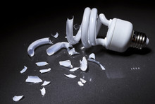 A Broken Compact Fluorescent CFL Bulb Lays Shattered In Shards Of Glass On A Hard Surface With A Dark Background