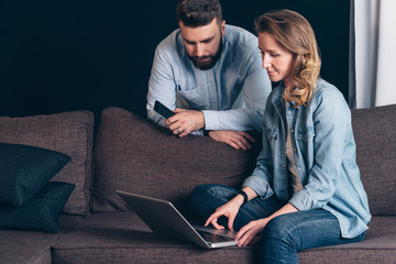 Wall Mural - Young woman in denim shirt,sitting at home on couch and using laptop.Guy holding smartphone.Couple shopping online,surfing internet.