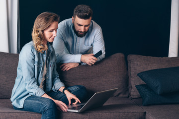 Young woman in denim shirt,sitting at home on couch and using laptop.Nearby stands man and looking on computer screen.Guy holding smartphone.Couple shopping online,surfing internet. Freelancers works.