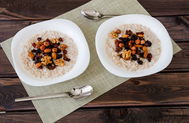 Two plates of homemade oatmeal porridge with dried plums, almonds, hazelnuts, walnuts and dates on old dark grey rustic wooden table with spoon. Hot and healthy food for Breakfast.
