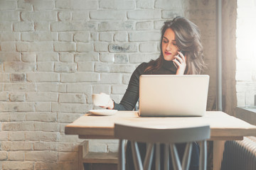 Wall Mural - Front view of young businesswoman sitting at table in coffee shop and talking on cell phone while drinking coffee and using laptop. In background white brick wall. Freelancer working outside office.