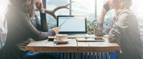 Teamwork, two young businesswomen sitting across table from each other. On table laptop, coffee cup and tablet computer. First girl showing pencil on computer screen, other is talking on cell phone.
