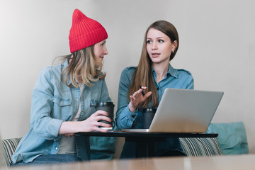 Wall Mural - Teamwork, two young businesswomen sitting at table in cafe, drinking coffee and working. On table cups of coffee and laptop. Students are learning online. Meeting of two friends in a coffee house.