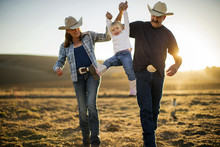 Farmer And His Wife Swinging Their Daughter Into The Air Between Them On Their Ranch At Sunset.