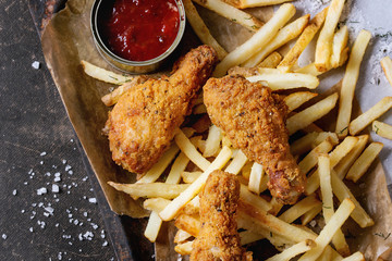 Fast food fried crispy chicken legs and french fries potatoes with salt and ketchup sauce served on baking paper in old rusty oven tray over dark texture background. Top view, space for text