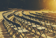 Empty Seat Rows Of Folding Chairs On Ground Before A Event Parallel And Rounded Arranged, Multiple Black Chairs On Street On Sunny Day In Park With People On Grass Meadow In Distance