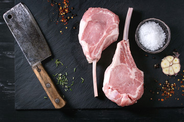 Two raw uncooked Veal tomahawk steak with garlic, salt, seasoning and vintage butcher cleaver on black stone slate board. Top view with space.