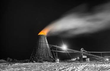 A large coned industrial wood chip burner with a fire inside shooting sparks out to the top in orange selective color at a sawmill yardsite in black and white at night in winter