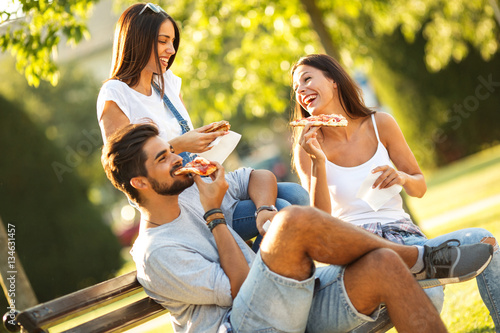 Group Of Young Friends Sitting On Park Bench And Eating Pizza Fast