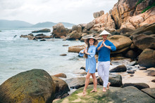 Young Couple In Love, Dressed Blue Shirt And Vietnamese Hats In Yoga Pose, Holding Hands On The Beach, Looking At The Sea. Big Stones Background. Concept Of Family