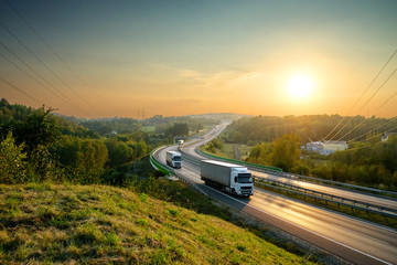 Wall Mural - white trucks driving on the highway winding through forested landscape at sunset.
