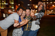© AYAimages - Multi-ethnic millenial group of friends taking a selfie photo with mobile phone on rooftop terrasse using flash at night time