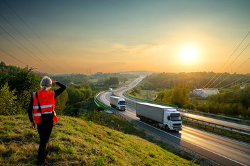 Wall Mural - the man in the orange safety vest watching white trucks driving on a highway in the landscape at sun