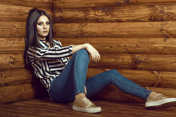 Portrait of young sexy dark-haired model wearing skinny high-waisted jeans, striped tied up shirt with pockets, choker and golden sneakers sitting with her legs spread on the wooden floor in log cabin