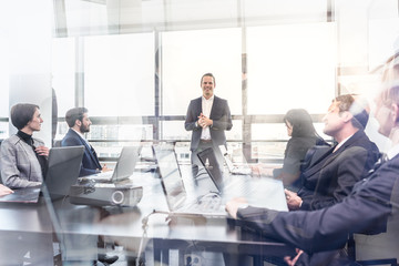 Wall Mural - Successful team leader and business owner leading informal in-house business meeting. Business people working on laptops in foreground and glass reflections. Business and entrepreneurship concept.