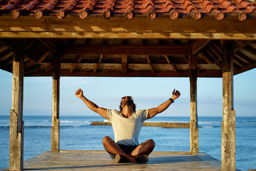 Wall Mural - Joy and freedom. Happy young african man rising hands up while sitting on deck near the sea.