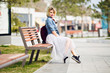 Young cute blond girl with short hair and bright pink lips sitting on a wooden bench and smiling wearing denim blue shirt, grey tulle skirt, black sneakers and marsala backpack