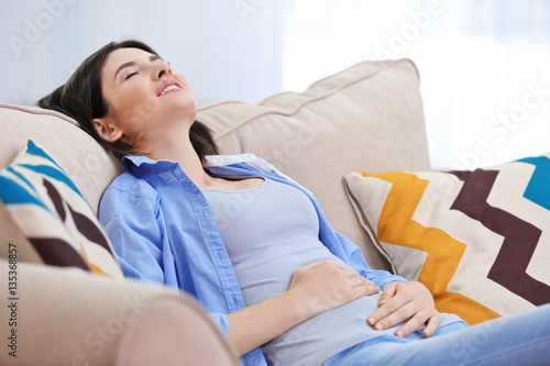 Fotografia, Obraz Beautiful young woman suffering from abdominal pain while sitting on sofa at hom
