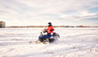 A teenage girl in bright colored winter gear and a helmet snowmobiling across a snow covered field with a farmyard in the background in a winter landscape
