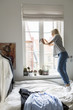 © Maskot - Full length of teenage girl looking through window while standing on bed at home