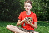 Fototapeta Londyn - Cute teenager boy wearing red t-shirt sitting on a lawn in a summer garden holding turtle looking at camera smiling